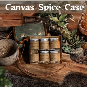 May include: A canvas spice case with six glass spice jars, each wrapped in tan fabric. The jars have silver lids and are held in place by a brown leather case. The text "Canvas Spice Case" is at the top of the image. A small green leather pouch is also visible.