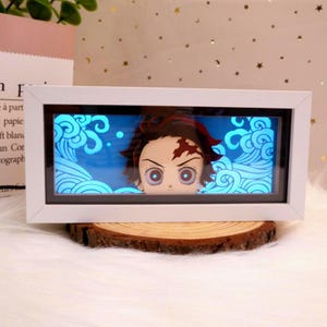 May include: A white framed LED light box with an anime character design. The character has red hair and a forehead scar. The background is blue with a wave pattern. The light box is on a wooden slice.
