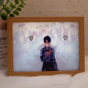 Peut inclure: Un cadre photo rectangulaire en bois avec une image imprimée d'un personnage d'anime. L'image présente un personnage en uniforme sombre avec un groupe de personnages flous en arrière-plan. Le cadre est de couleur marron clair.