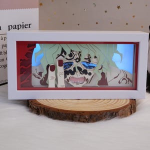 May include: A white framed artwork with a layered paper illustration of a character with green hair and blue eyes. The character is making a rude gesture with their fingers. The artwork is set against a red background.