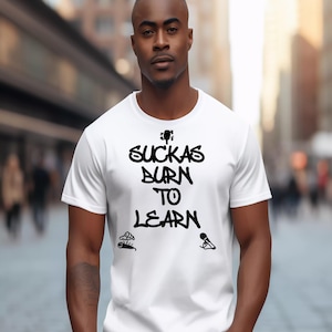 Op de afbeelding: Wit T-shirt met korte mouwen met de zwarte tekst "Suckas Burn to Learn" en kleine illustraties. De persoon draagt het T-shirt buiten, met een vage achtergrond.
