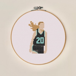 May include: A cross-stitch hoop featuring a basketball player in a black and teal jersey with the number 20. The player's hair is light brown, and the background is white. The hoop is made of wood and has a metal clasp.