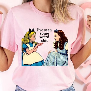 May include: Pink t-shirt featuring a graphic of two Alice in Wonderland characters with the text "I've seen some weird shit." The illustration depicts the characters in blue dresses and white aprons, with one blonde and one brunette.