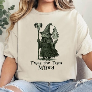 May include: Beige t-shirt featuring a whimsical illustration of a frog wizard in a black robe and hat, holding a staff and a flame. The text "T'was the 'Tism M'Lord" is printed below the image.