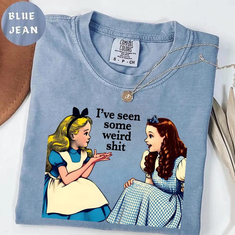 May include: A light blue t-shirt with the text "I've seen some weird shit" and an illustration of two girls in dresses. The shirt has a Comfort Colors tag and a silver necklace. The shirt is labeled "Blue Jean".