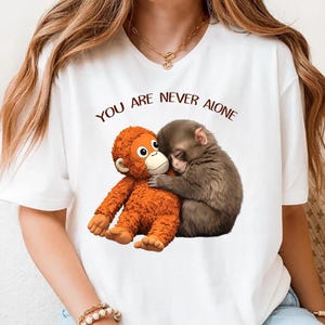 May include: White t-shirt featuring a graphic of two monkeys hugging. One is orange, the other is brown. The text "YOU ARE NEVER ALONE" is printed above the monkeys in brown letters. The shirt has a classic crew neck.