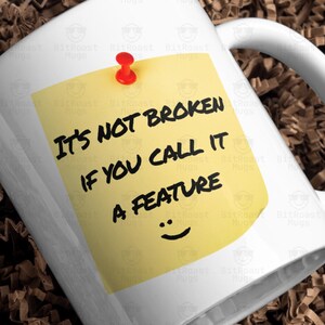 May include: White ceramic mug with a yellow sticky note pinned to it with a red push pin. The note reads "It's NOT BROKEN IF YOU CALL IT A FEATURE" with a smiley face. The mug is in a white box filled with brown paper.