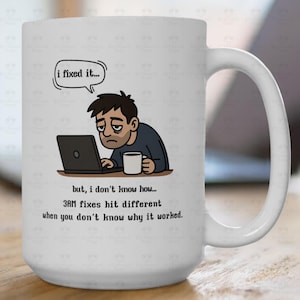 May include: White ceramic mug with a humorous cartoon of a person looking tired while working on a laptop. The mug has the text "i fixed it..." and "3AM fixes hit different when you don't know why it worked."