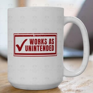 May include: White ceramic mug with a red rectangular graphic that reads "WORKS AS UNINTENDED" with a checkmark. The mug is on a wooden surface with a laptop in the background.