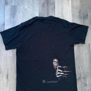 May include: Black t-shirt featuring a graphic of a creature's eye and clawed hand emerging from the darkness. The design includes a PlayStation logo and text. The shirt is made of a soft material.