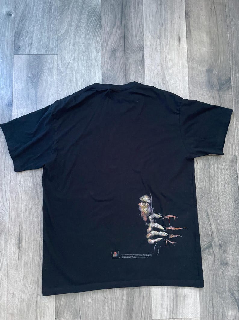 May include: Black t-shirt featuring a graphic of a creature's eye and clawed hand emerging from the darkness. The design includes a PlayStation logo and text. The shirt is made of a soft material.