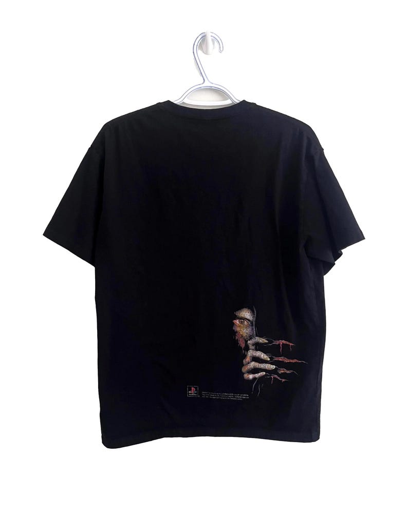 May include: Black t-shirt with a graphic of a face and hand with long, sharp nails. The graphic is on the lower right side of the shirt. The shirt is hanging on a silver hanger.