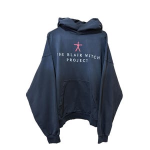 May include: Dark navy blue hoodie with a front pocket and a hood. The text "THE BLAIR WITCH PROJECT" is printed in white, with a pink symbol above the text. The hoodie has long sleeves and ribbed cuffs and hem.