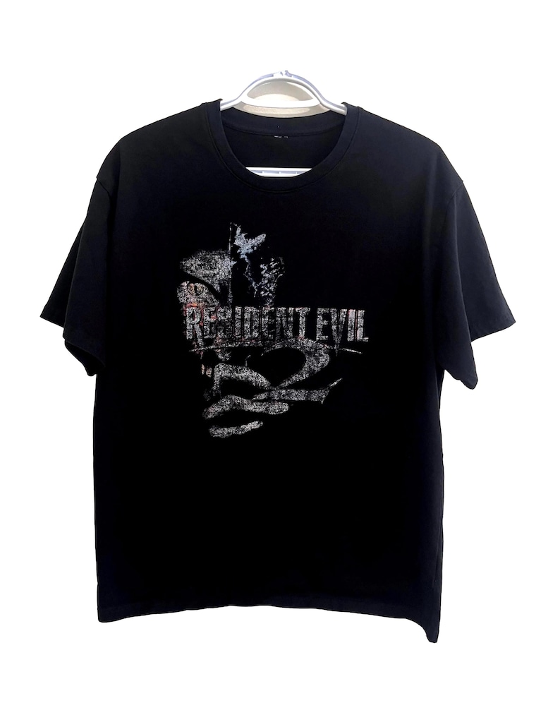 May include: Black t-shirt featuring the text "RESIDENT EVIL 2" in a distressed, white font. The graphic includes a stylized image. The shirt is hanging on a white hanger.