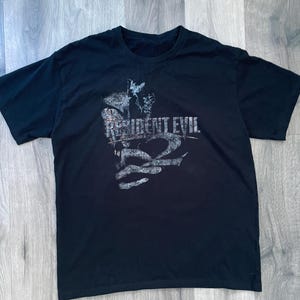 May include: Black t-shirt featuring the text "RESIDENT EVIL 2" in a distressed, gray font. The graphic includes a stylized image. The shirt is made of a soft material and has a crew neck.