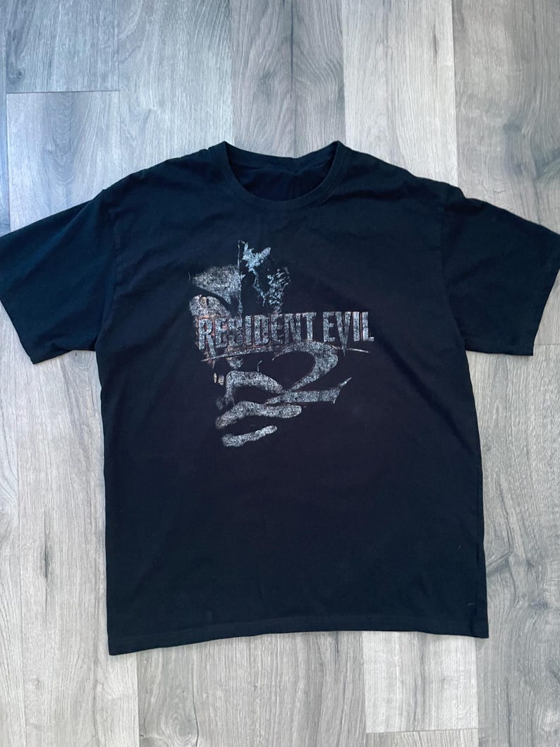 May include: Black t-shirt featuring the text "RESIDENT EVIL 2" in a distressed, gray font. The graphic includes a stylized image. The shirt is made of a soft material and has a crew neck.
