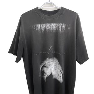 May include: Dark gray t-shirt featuring a forest graphic and the text "THE BLAIR WITCH PROJECT". The shirt has a distressed look and a crew neck.