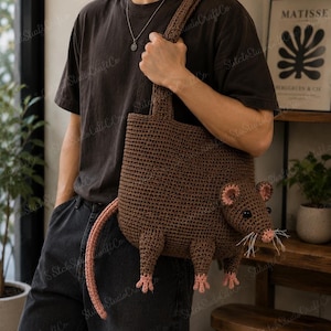 May include: A brown crochet tote bag shaped like a rat, featuring a long tail, four small legs, pink-accented ears and nose, black button eyes, and white thread whiskers. The bag has a single shoulder strap and a textured, hand-stitched appearance.