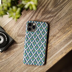 May include: A patterned phone case with a repeating design in blue, yellow, and teal. The case is on a wooden surface, with a camera and greenery in the background. The case has a geometric pattern.