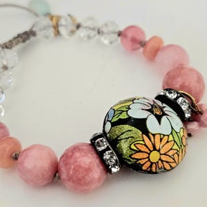May include: A pink and white beaded bracelet with a large, round, black bead featuring a colorful floral design. The bead is accented with clear rhinestones.