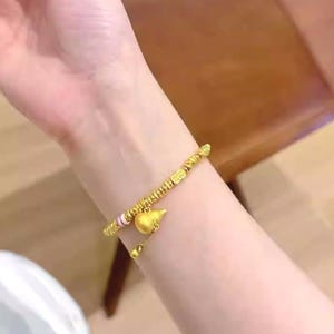 May include: A gold-colored bracelet with a gourd-shaped charm and small beads. The bracelet is worn on a wrist, and the charm hangs below the wrist. The bracelet has a delicate design and appears to be made of metal.