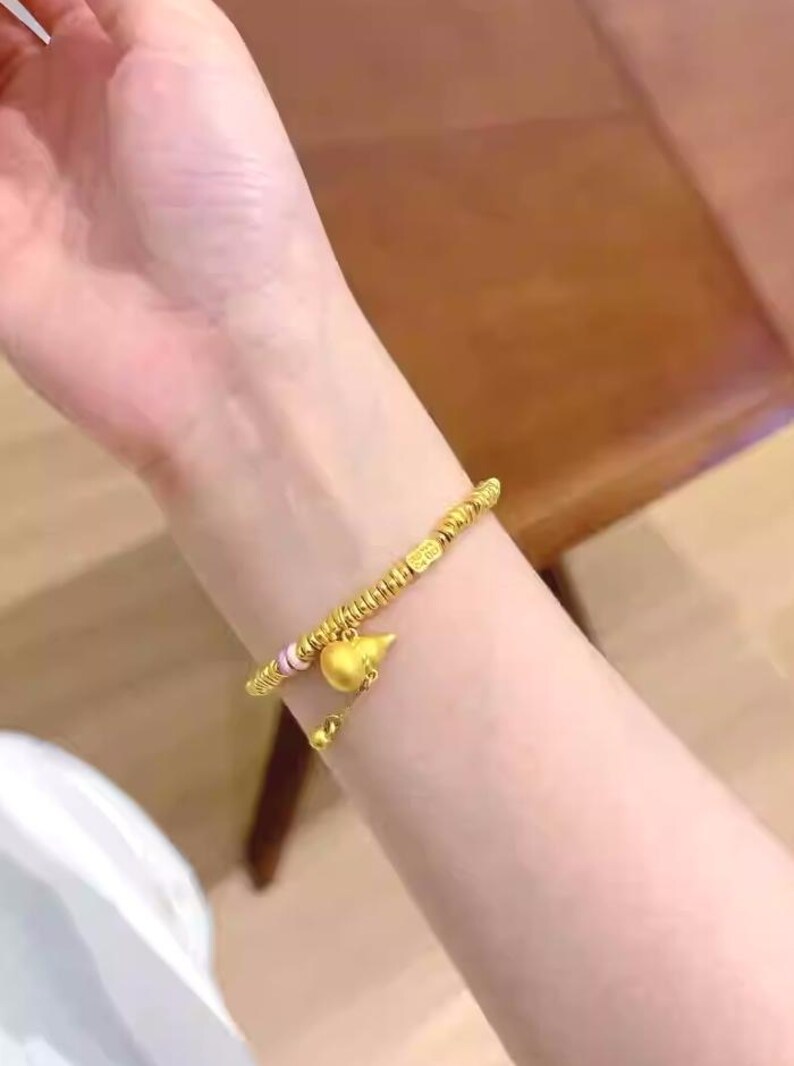 May include: A gold-colored bracelet with a gourd-shaped charm and small beads. The bracelet is worn on a wrist, and the charm hangs below the wrist. The bracelet has a delicate design and appears to be made of metal.