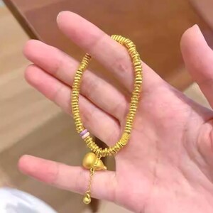 May include: Gold-colored bracelet with a series of small, round beads. The bracelet has a charm of a small, gold-colored gourd with a dangling gold bead. The bracelet is held in a hand.