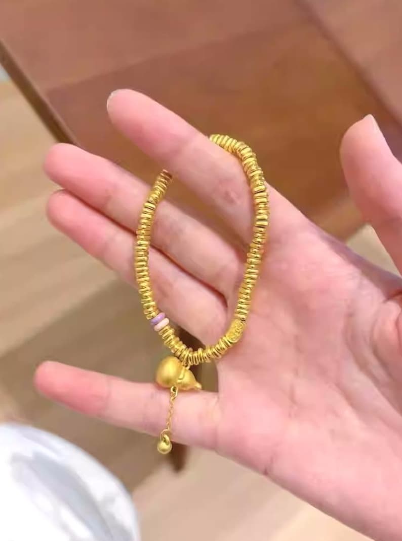May include: Gold-colored bracelet with a series of small, round beads. The bracelet has a charm of a small, gold-colored gourd with a dangling gold bead. The bracelet is held in a hand.