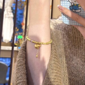 May include: Gold-toned bracelet featuring a beaded design with a small charm and a dangling pendant. The bracelet has a delicate appearance and is worn on the wrist. The charm has a unique shape, and the pendant adds a touch of elegance.