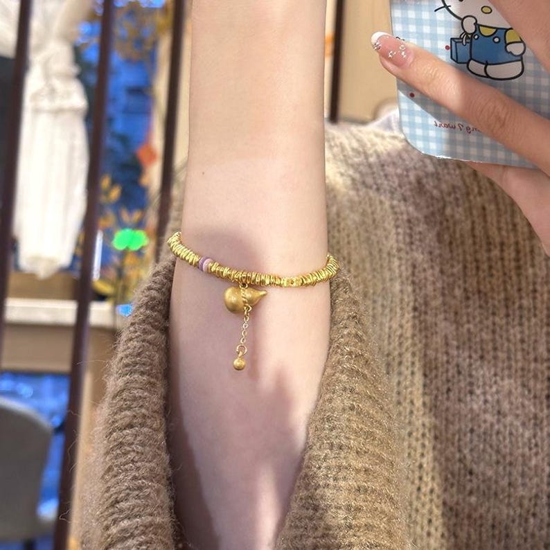 May include: Gold-toned bracelet featuring a beaded design with a small charm and a dangling pendant. The bracelet has a delicate appearance and is worn on the wrist. The charm has a unique shape, and the pendant adds a touch of elegance.
