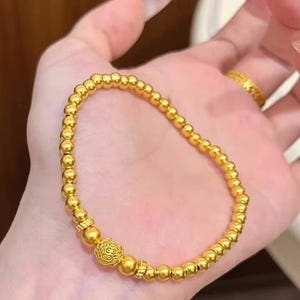 K&ouml;nnte beinhalten: Ein goldfarbenes Perlenarmband mit einem dekorativen kugelf&ouml;rmigen Anh&auml;nger. Das Armband besteht aus kleinen, runden Perlen und hat einen detaillierten Anh&auml;nger. Das Armband wird auf einer Hand gezeigt.