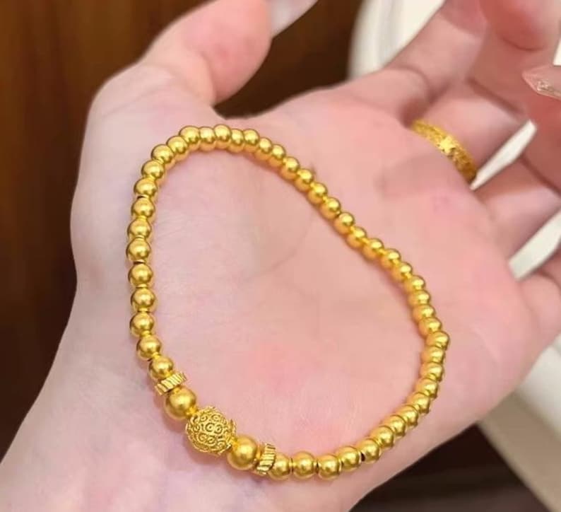 K&ouml;nnte beinhalten: Ein goldfarbenes Perlenarmband mit einem dekorativen kugelf&ouml;rmigen Anh&auml;nger. Das Armband besteht aus kleinen, runden Perlen und hat einen detaillierten Anh&auml;nger. Das Armband wird auf einer Hand gezeigt.