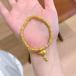 May include: A gold-colored bracelet with a beaded design and a charm. The bracelet is held in a hand. The charm is a small bird-like shape with a dangling bead. The bracelet appears to be made of metal.