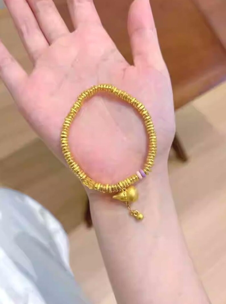 May include: A gold-colored bracelet with a beaded design and a charm. The bracelet is held in a hand. The charm is a small bird-like shape with a dangling bead. The bracelet appears to be made of metal.
