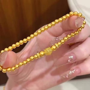 K&ouml;nnte beinhalten: Goldenes Perlenarmband mit einer dekorativen Filigranperle und zwei klaren, oval geformten Steinen. Das Armband wird in einer Hand mit langen, manik&uuml;rten N&auml;geln gehalten.