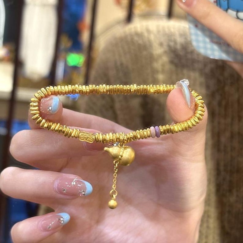 May include: A gold-colored bracelet with a textured, coiled design. The bracelet features a small, gold-colored teapot charm hanging from a delicate chain. The bracelet is held up by a hand with manicured nails.