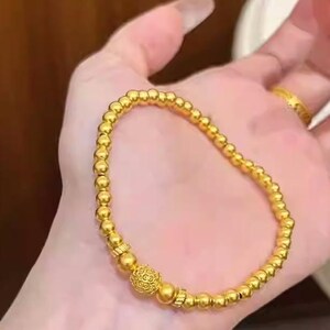 K&ouml;nnte beinhalten: Ein goldfarbenes Perlenarmband mit einer dekorativen Mittelperle. Das Armband besteht aus kleinen, runden, goldfarbenen Perlen, die aneinandergereiht sind. Die Mittelperle hat ein aufwendiges Design. Das Armband wird auf einer Hand gezeigt.