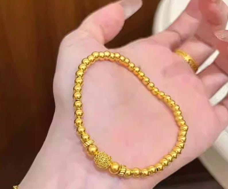 K&ouml;nnte beinhalten: Ein goldfarbenes Perlenarmband mit einer dekorativen Mittelperle. Das Armband besteht aus kleinen, runden, goldfarbenen Perlen, die aneinandergereiht sind. Die Mittelperle hat ein aufwendiges Design. Das Armband wird auf einer Hand gezeigt.