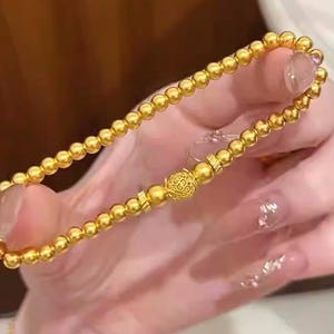 K&ouml;nnte beinhalten: Ein goldfarbenes Perlenarmband mit einem dekorativen Mittelst&uuml;ck. Das Armband besteht aus kleinen, runden Perlen und einem detaillierten, verzierten Mittelst&uuml;ck. Das Armband wird vor einem unscharfen Hintergrund gehalten.