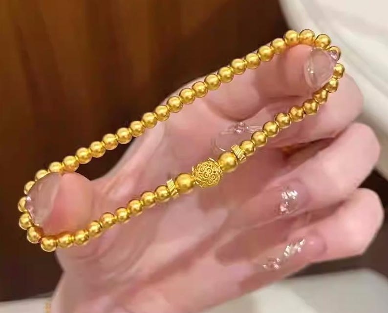 K&ouml;nnte beinhalten: Ein goldfarbenes Perlenarmband mit einem dekorativen Mittelst&uuml;ck. Das Armband besteht aus kleinen, runden Perlen und einem detaillierten, verzierten Mittelst&uuml;ck. Das Armband wird vor einem unscharfen Hintergrund gehalten.