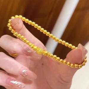 K&ouml;nnte beinhalten: Ein goldfarbenes Armband mit Perlen und einer dekorativen Mittelperle. Das Armband besteht aus kleinen, runden Perlen und einer gr&ouml;&szlig;eren, verzierten Perle. Das Armband wird gezeigt, um sein Design und seine Farbe zu pr&auml;sentieren.