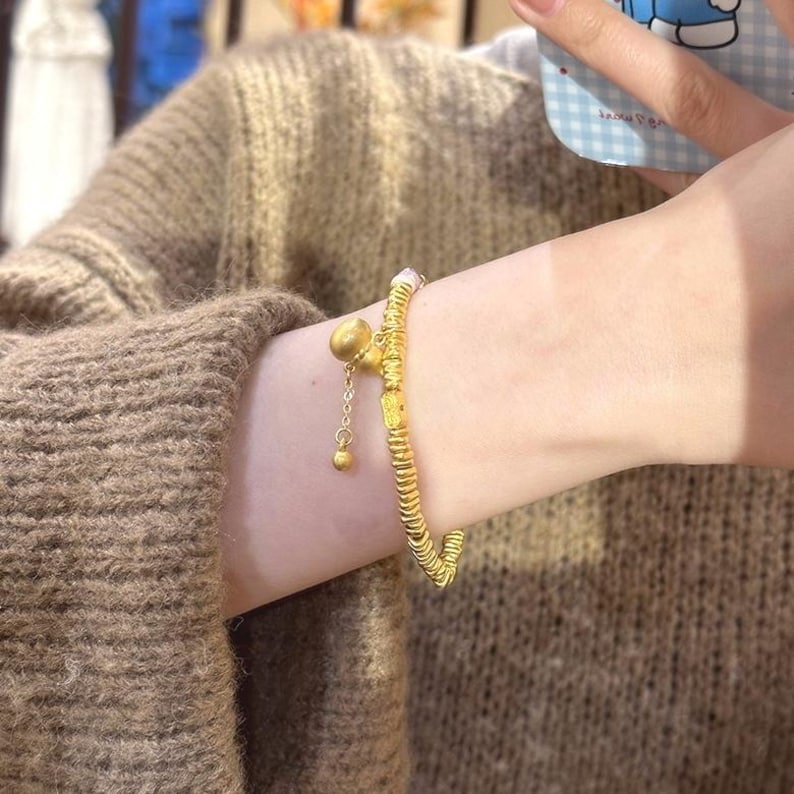 May include: A gold-colored bracelet with a bell charm and small dangling ball. The bracelet is worn on a wrist, and the design features a series of small, connected segments. The bell charm hangs from a delicate chain.