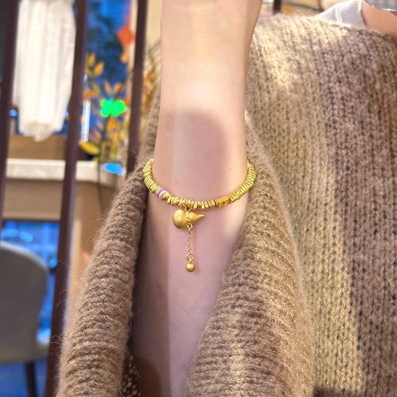 May include: Gold-toned bracelet featuring a charm with a bird-like design and a dangling bead. The bracelet has a textured, segmented band with small colorful accents. The jewelry is displayed on a person wearing a brown sweater.