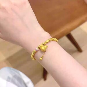 May include: Gold-colored bracelet with a gourd-shaped charm and a small dangling bead. The bracelet has a beaded design with a section of pink and white beads. The bracelet is worn on a wrist.