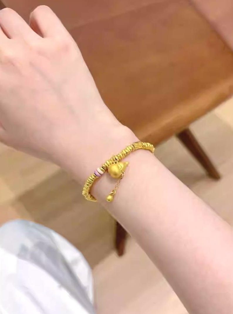 May include: Gold-colored bracelet with a gourd-shaped charm and a small dangling bead. The bracelet has a beaded design with a section of pink and white beads. The bracelet is worn on a wrist.
