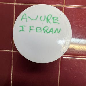 Puede incluir: Un objeto blanco y circular con el texto manuscrito "AWURE I FERAN" en verde. El objeto está sobre un fondo de baldosas de color rojo oscuro.