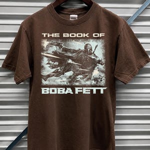 May include: Dark chocolate brown t-shirt featuring a graphic of Boba Fett from "The Book of Boba Fett". The design shows Boba Fett in action with a blaster. The text "THE BOOK OF" and "BOBA FETT" are printed in white.