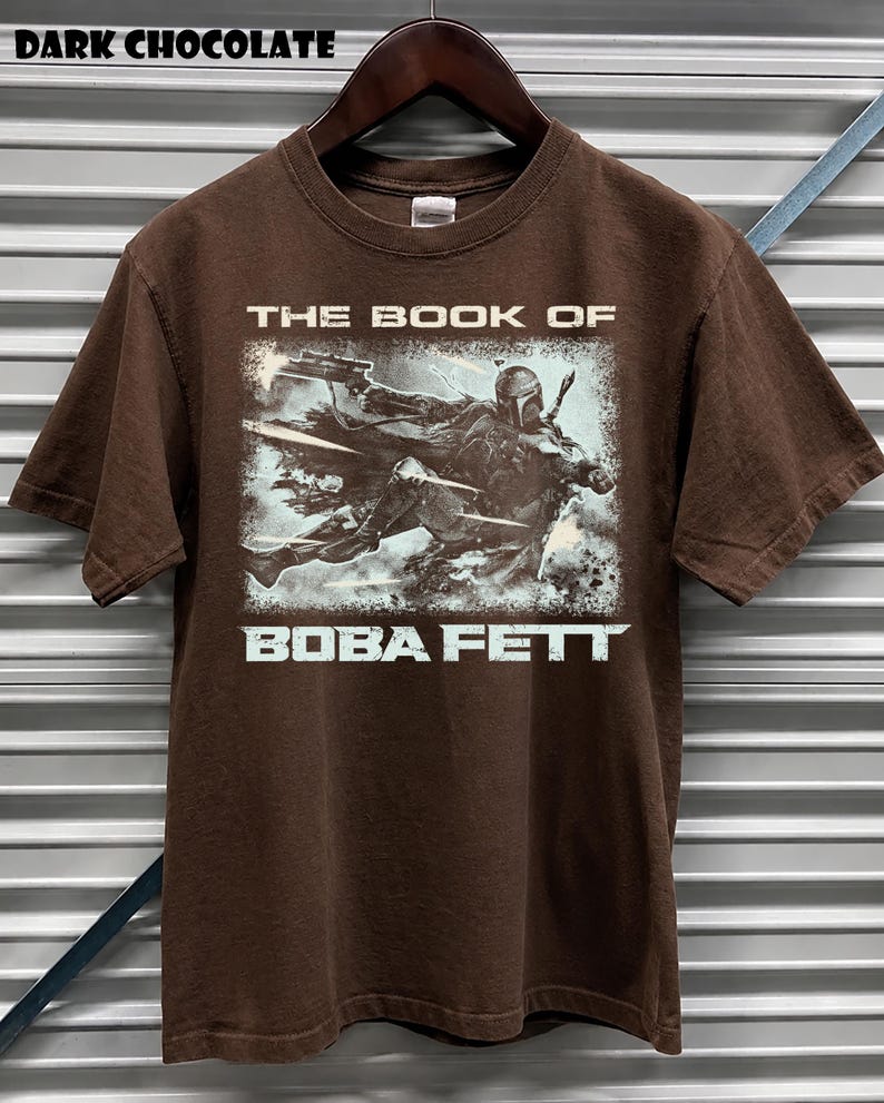 May include: Dark chocolate brown t-shirt featuring a graphic of Boba Fett from "The Book of Boba Fett". The design shows Boba Fett in action with a blaster. The text "THE BOOK OF" and "BOBA FETT" are printed in white.