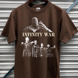 K&ouml;nnte beinhalten: Dunkel schokoladenbraunes T-Shirt mit einer Grafik aus dem Film "Infinity War". Das Design zeigt Thanos und andere Charaktere. Der Text "DARK CHOCOLATE" steht oben. Das T-Shirt h&auml;ngt an einem Holzkleiderb&uuml;gel.