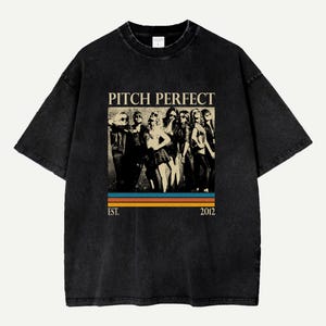 Puede incluir: Camiseta negra con un gráfico de un grupo de mujeres y el texto "PITCH PERFECT" sobre un diseño de rayas retro. El texto "EST. 2012" está debajo. La camiseta tiene un aspecto vintage y desgastado.